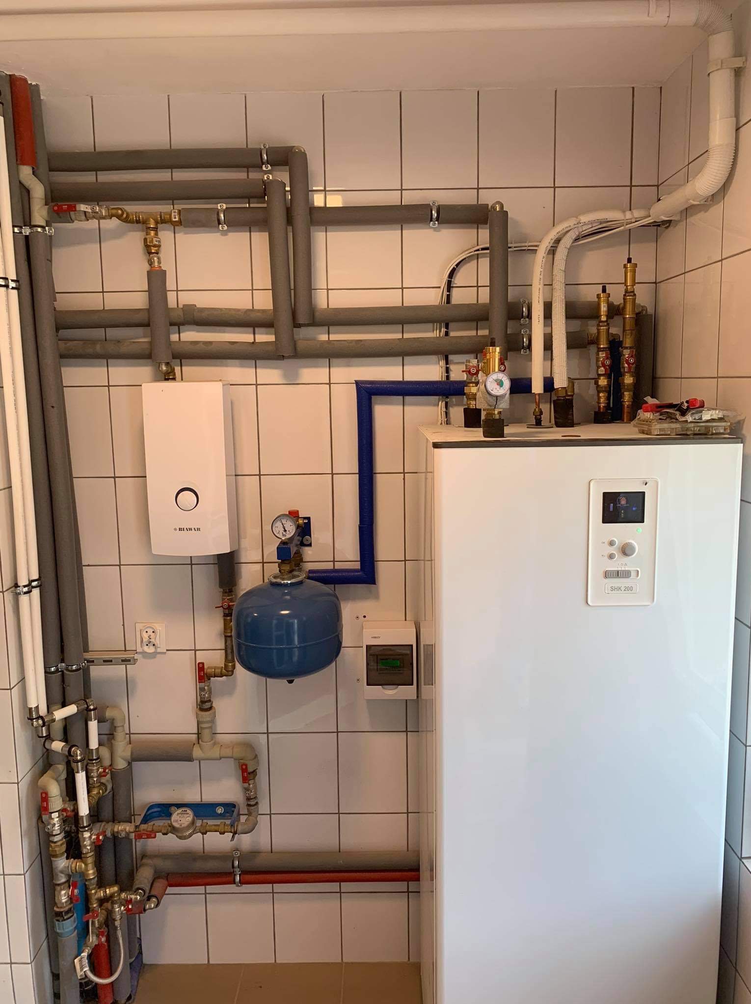 Biały zasobnik ciepłej wody użytkowej SHK 200 z orurowaniem, manometrem i zaworami na tle białych płytek. Widoczny też biały piec Biowar i niebieski zbiornik przeponowy.