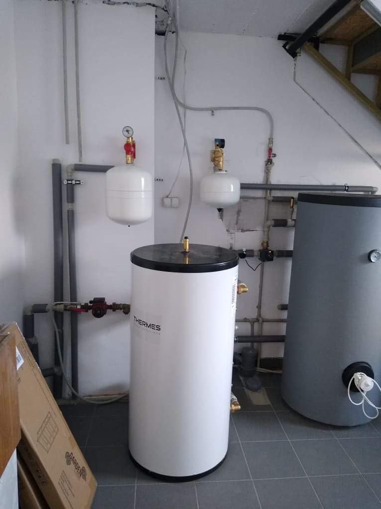 Biały bojler Thermes z podłączoną instalacją hydrauliczną i zbiornikiem wyrównawczym na tle szarej ściany i płytek. Widoczne rury i elementy instalacji.