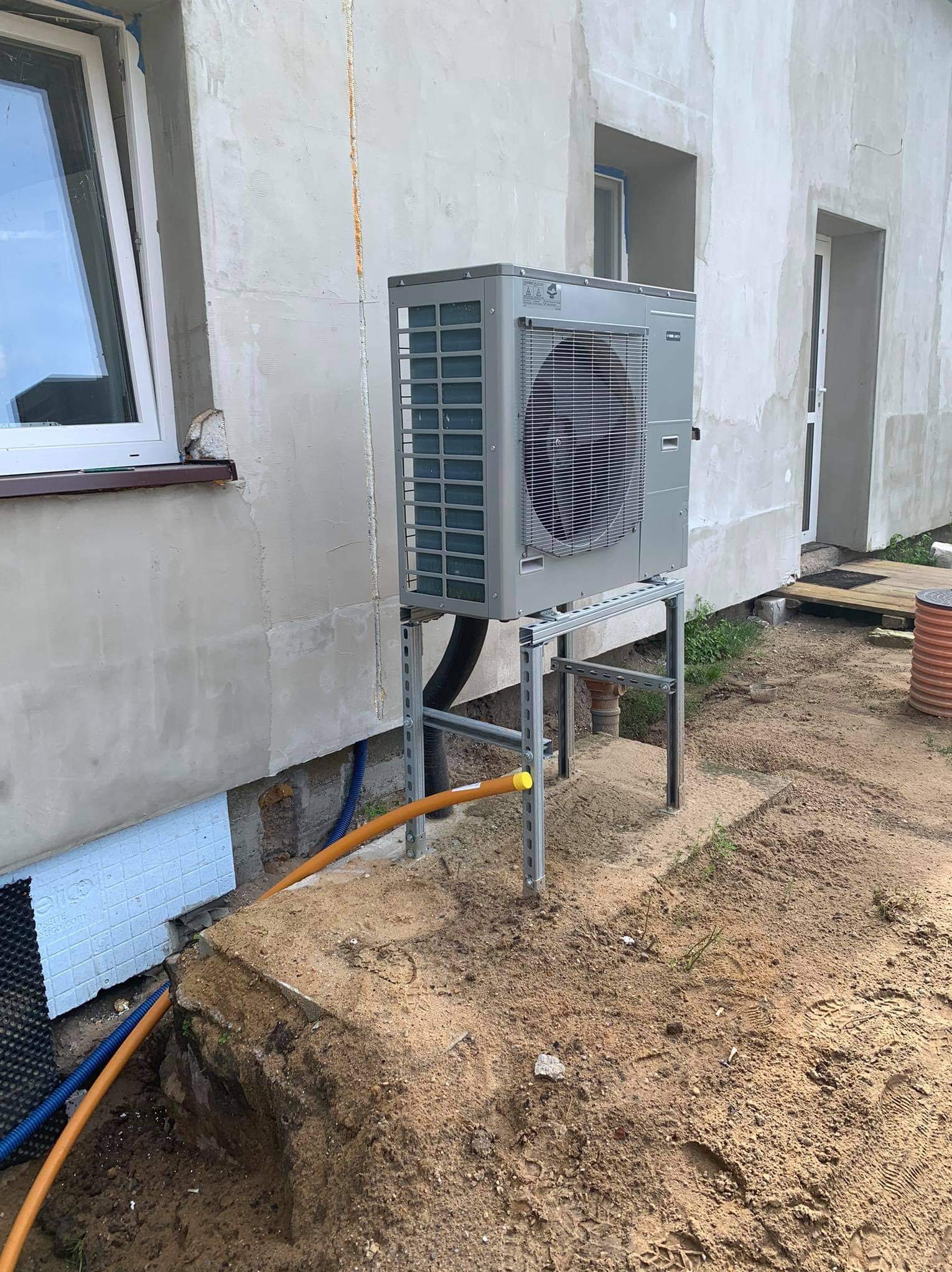 Szara pompa ciepła na metalowej konstrukcji, zamontowana przy ścianie budynku w trakcie prac ziemnych. Widoczne rury i elementy instalacji.