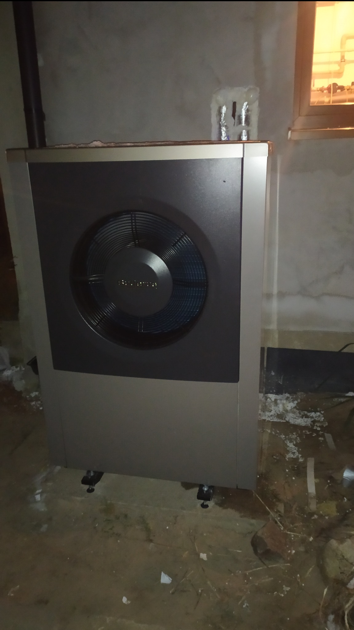 Szara pompa ciepła Buderus z widocznym wentylatorem na tle szarej ściany i okna. Widoczne ślady prac instalacyjnych na podłodze i ścianie.