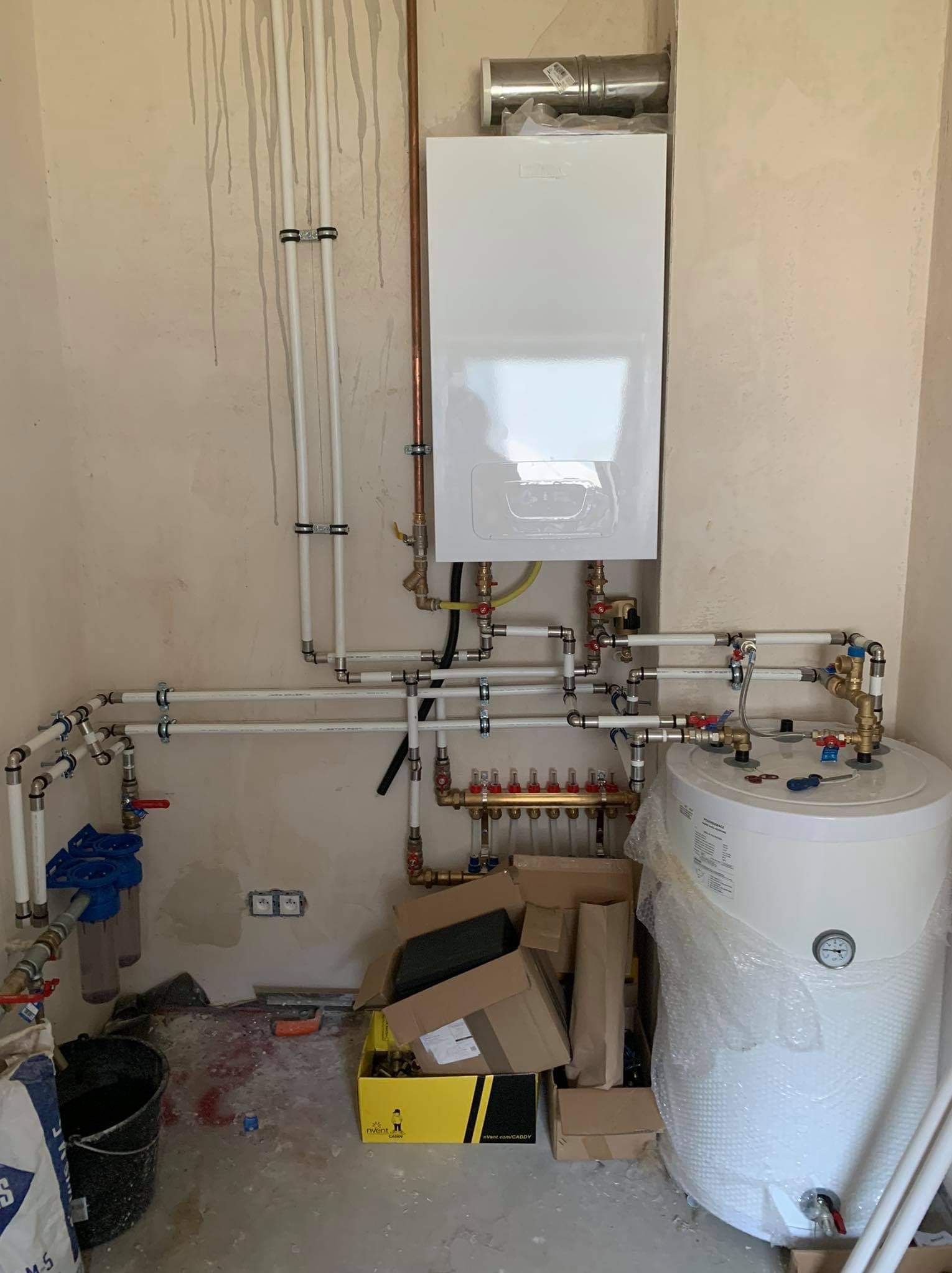 Biały piec gazowy zamontowany na ścianie, z podłączonymi rurami i zbiornikiem wody. Widoczne elementy instalacji hydraulicznej i gazowej w surowym wnętrzu.