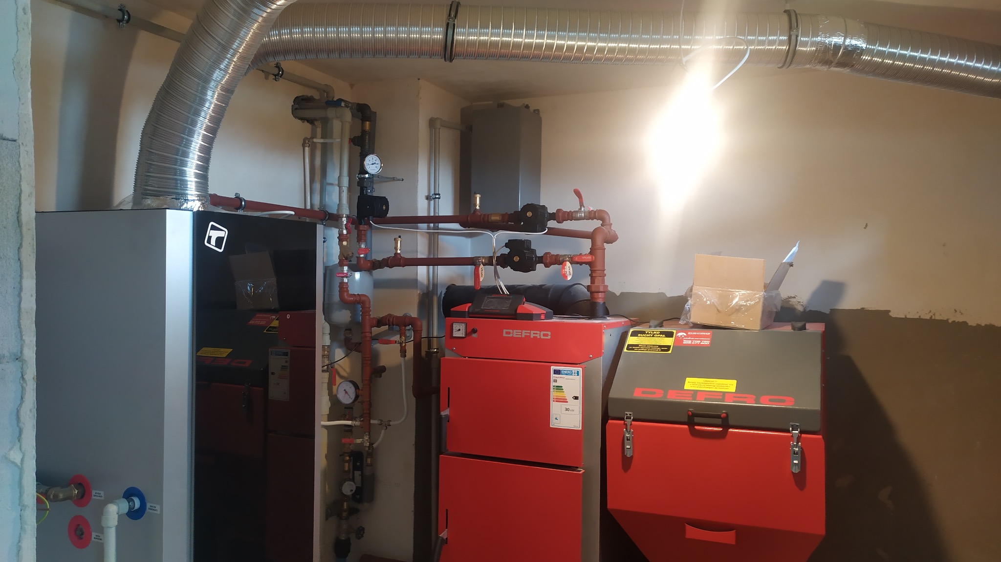 Nowoczesna kotłownia z piecami Defro, rurami i wentylacją. Widoczne elementy instalacji grzewczej i kominowe. Oświetlenie górne.