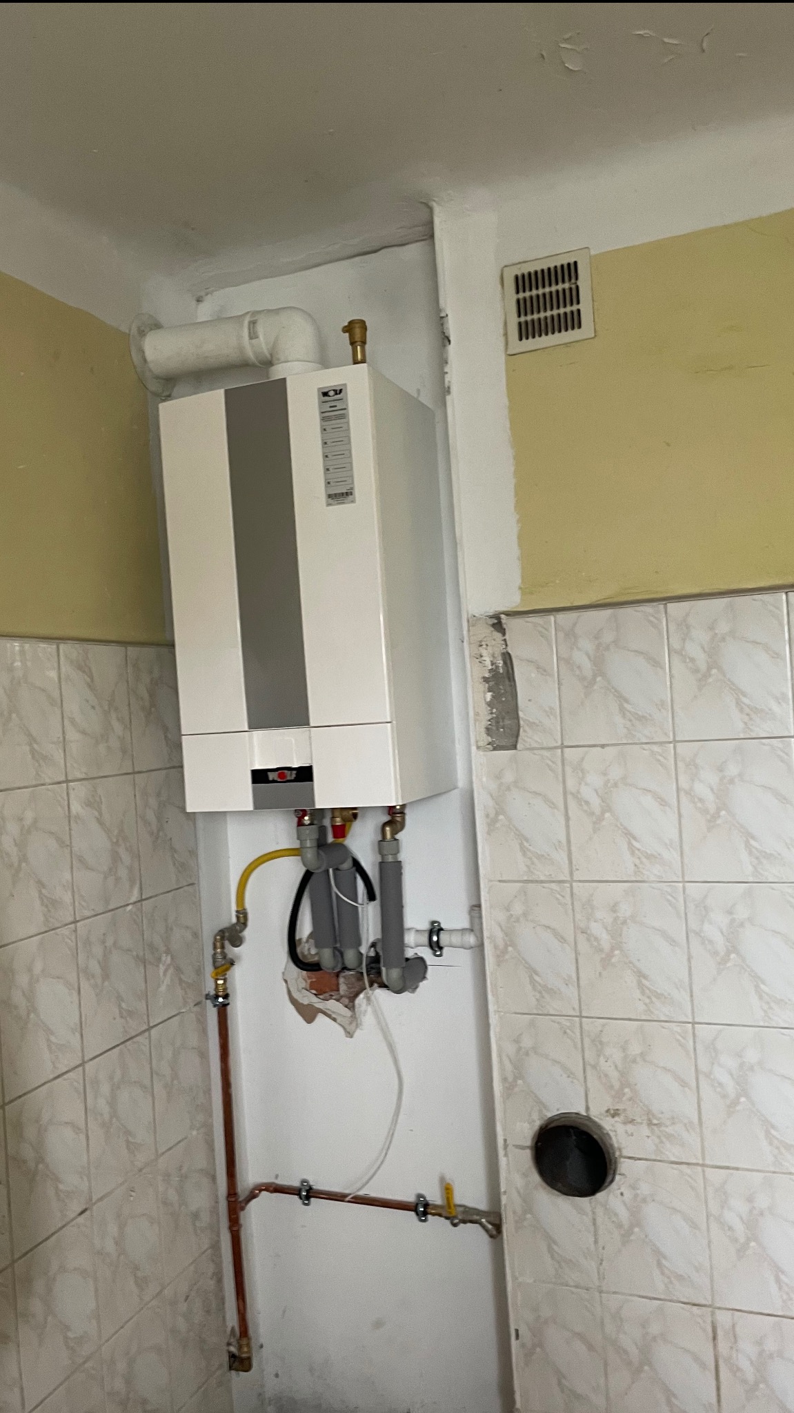 Montaż pieca gazowego Wolf w Przasnyszu. Widoczne rury i podłączenia. Biało-szary piec zawieszony na ścianie z jasnymi kafelkami.