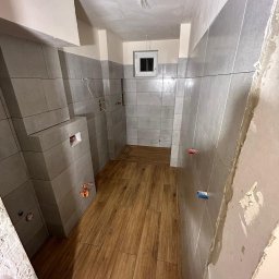 Dawid - Wnętrze łazienki w trakcie remontu: szare płytki ścienne, drewnopodobne panele podłogowe, przygotowane punkty instalacyjne, surowy tynk na suficie i ścianie po prawej.