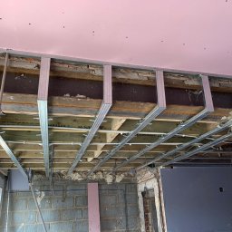 Dawid - Konstrukcja sufitu podwieszanego z profili stalowych, widoczne elementy instalacji elektrycznej i poziomica przytwierdzona do profilu, surowy stan wykończenia pomieszczenia.