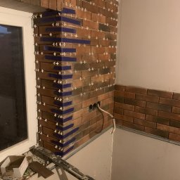 Dawid - Ściana wyłożona cegiełkami w trakcie prac, widoczne krzyżyki dystansowe i taśma malarska przy oknie, kabel elektryczny wystający ze ściany, bałagan na podłodze.