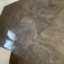 Dawid - Ułożone brązowo-szare płytki podłogowe z efektem marmuru, widoczne niedociągnięcia przy ścianie, odbicie światła na powierzchni płytek.