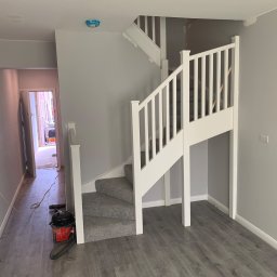 Dawid - Pomieszczenie po remoncie: szare ściany, białe schody z szarym dywanem, szara podłoga, widoczny odkurzacz i kabel na podłodze.