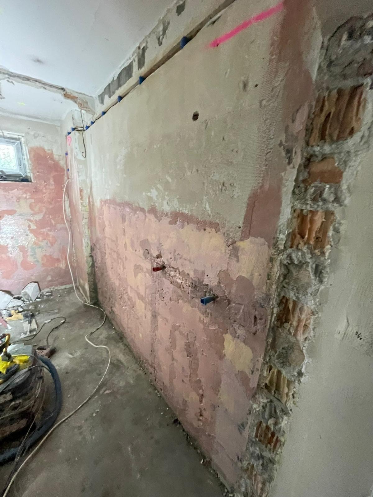 Ściany w trakcie przygotowania do remontu, widoczne fragmenty starej farby i tynku, odsłonięta cegła na rogu, instalacja wodna zabezpieczona korkami, różowe oznaczenia na ścianie.