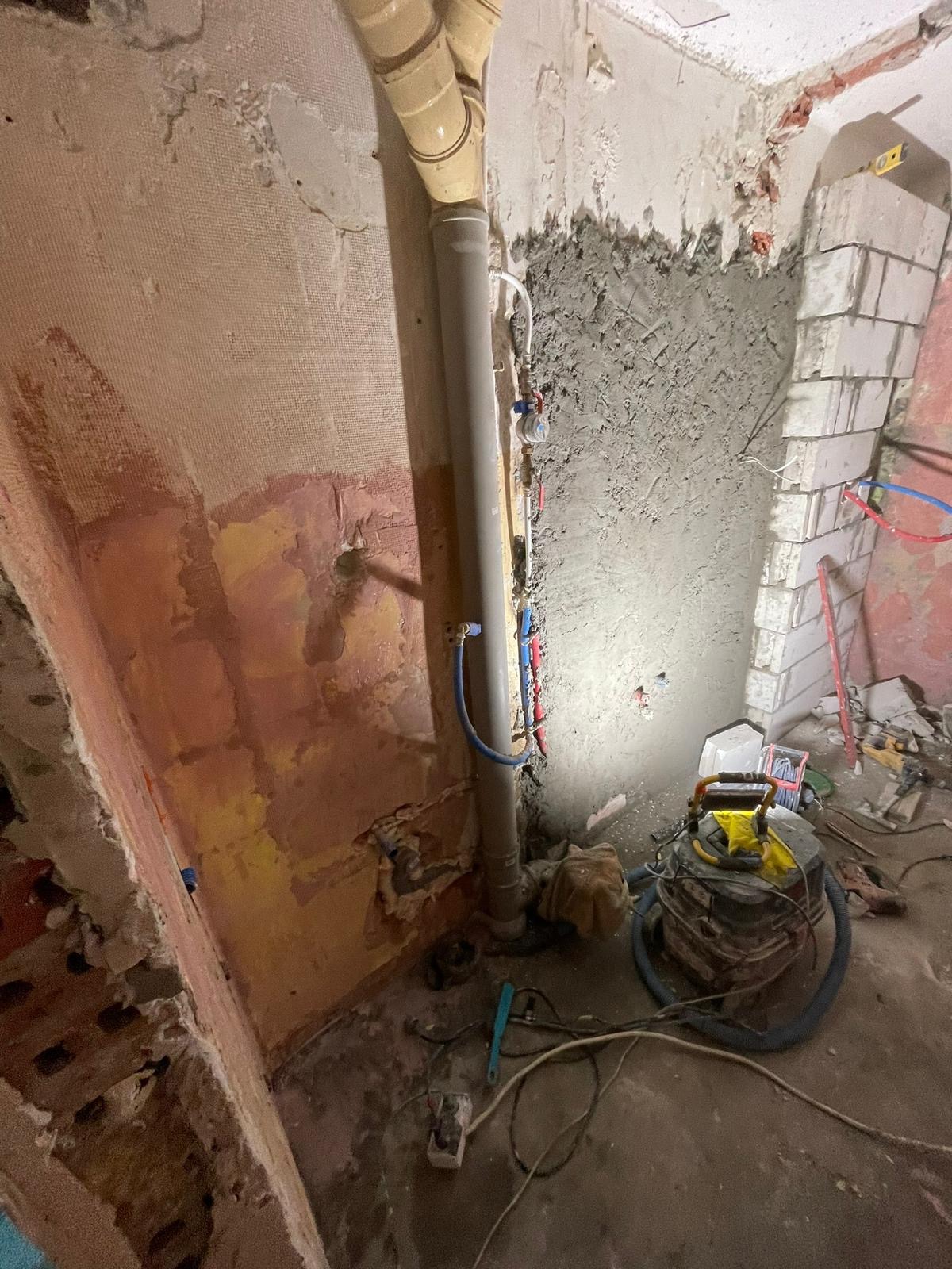 Wnętrze w trakcie remontu: ściana z resztkami starej farby i siatką, fragment ściany pokryty świeżym tynkiem, pionowe rury instalacyjne, narzędzia i odkurzacz przemysłowy na podłodze.