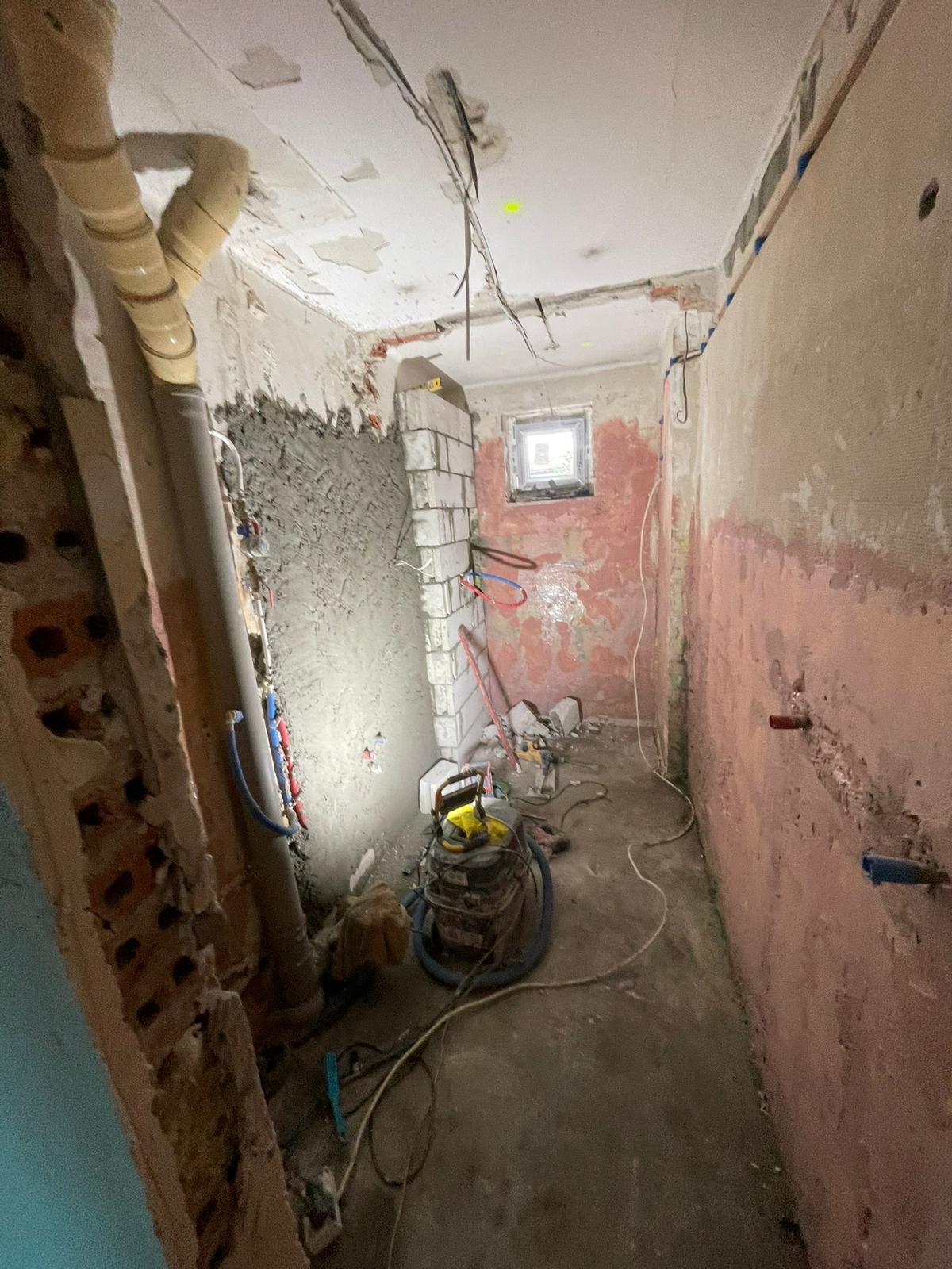 Łazienka w trakcie gruntownego remontu: odsłonięte instalacje, fragmenty cegieł, ślady po starych płytkach na ścianach, widoczne rury i kable, narzędzia budowlane leżące na podłodze, małe okno...