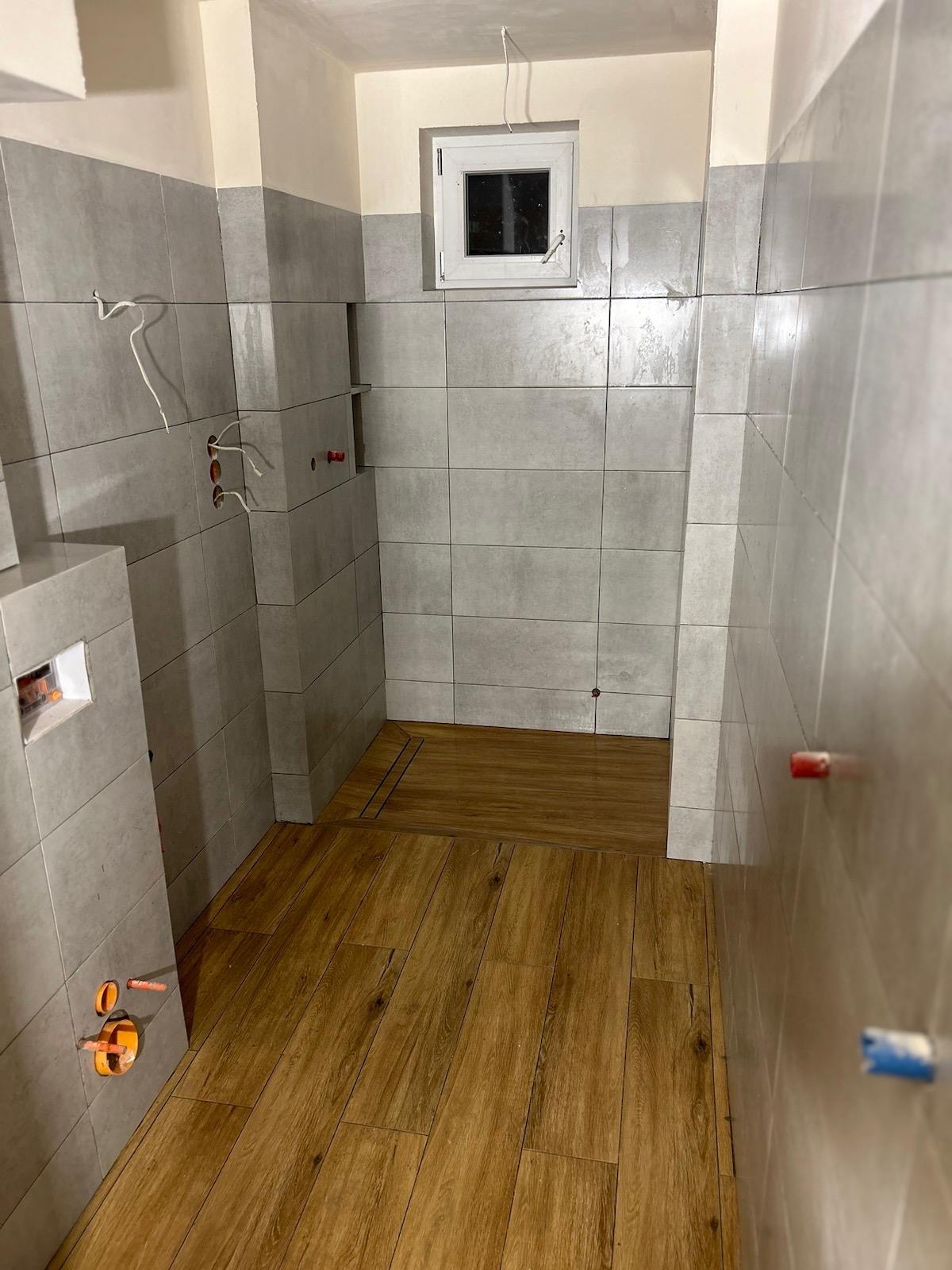 Wnętrze łazienki w trakcie remontu: szare płytki ścienne, drewnopodobna podłoga, widoczne instalacje hydrauliczne i elektryczne, małe okno. Częściowo wykończona kabina prysznicowa z odpływem liniowym.
