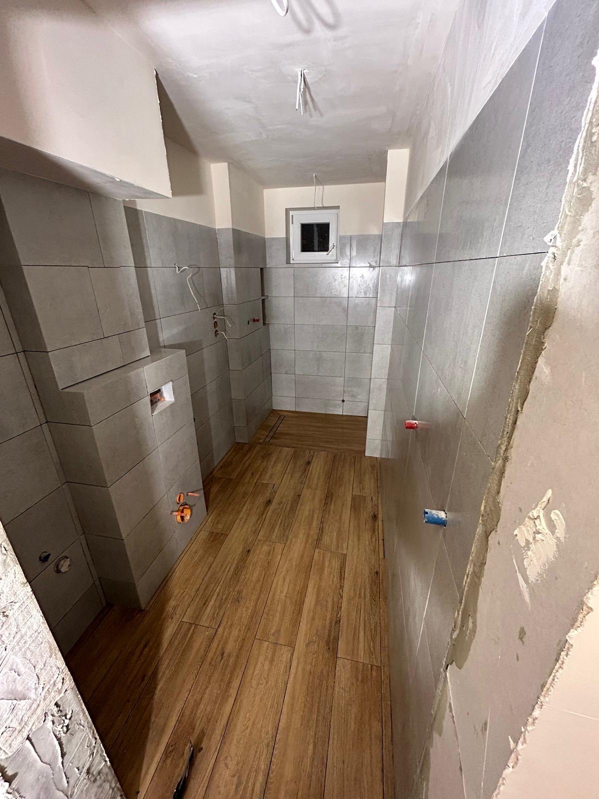 Wnętrze łazienki w trakcie remontu: szare płytki ścienne, drewnopodobne panele podłogowe, przygotowane punkty instalacyjne, surowy tynk na suficie i ścianie po prawej.