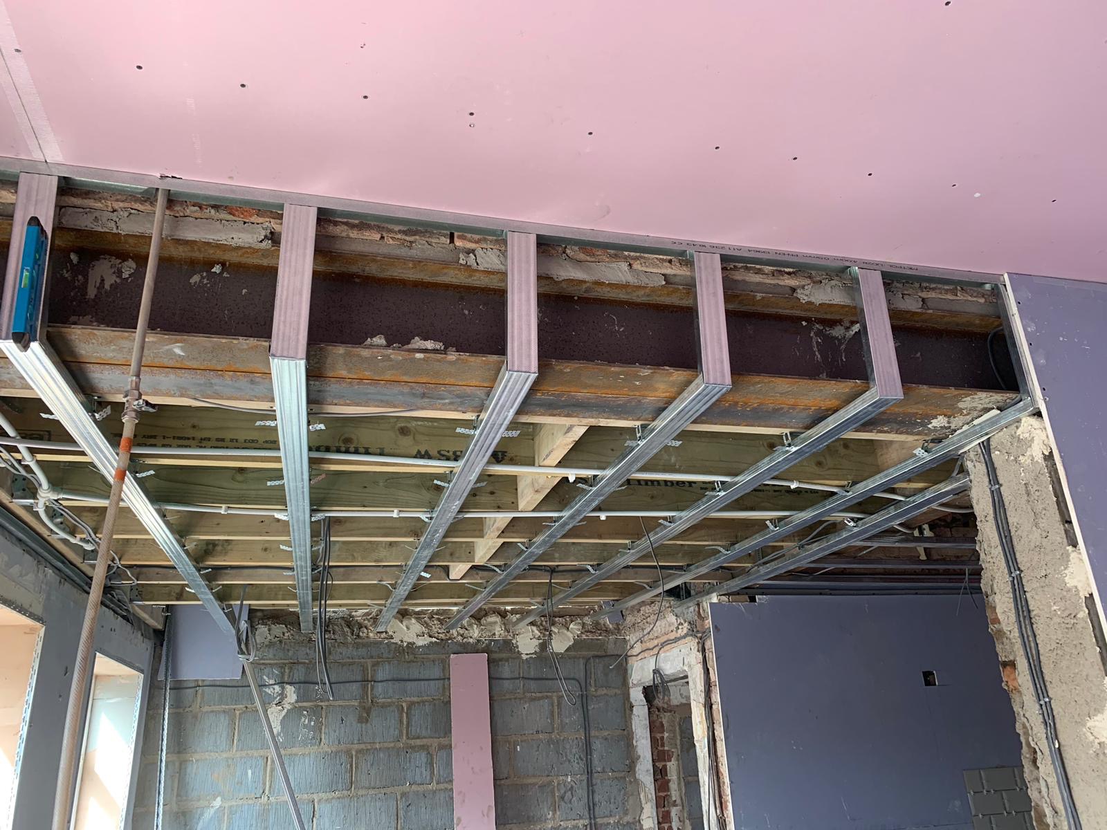 Konstrukcja sufitu podwieszanego z profili stalowych, widoczne elementy instalacji elektrycznej i poziomica przytwierdzona do profilu, surowy stan wykończenia pomieszczenia.