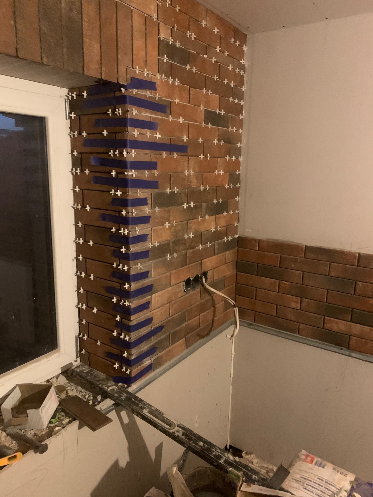 Ściana wyłożona cegiełkami w trakcie prac, widoczne krzyżyki dystansowe i taśma malarska przy oknie, kabel elektryczny wystający ze ściany, bałagan na podłodze.