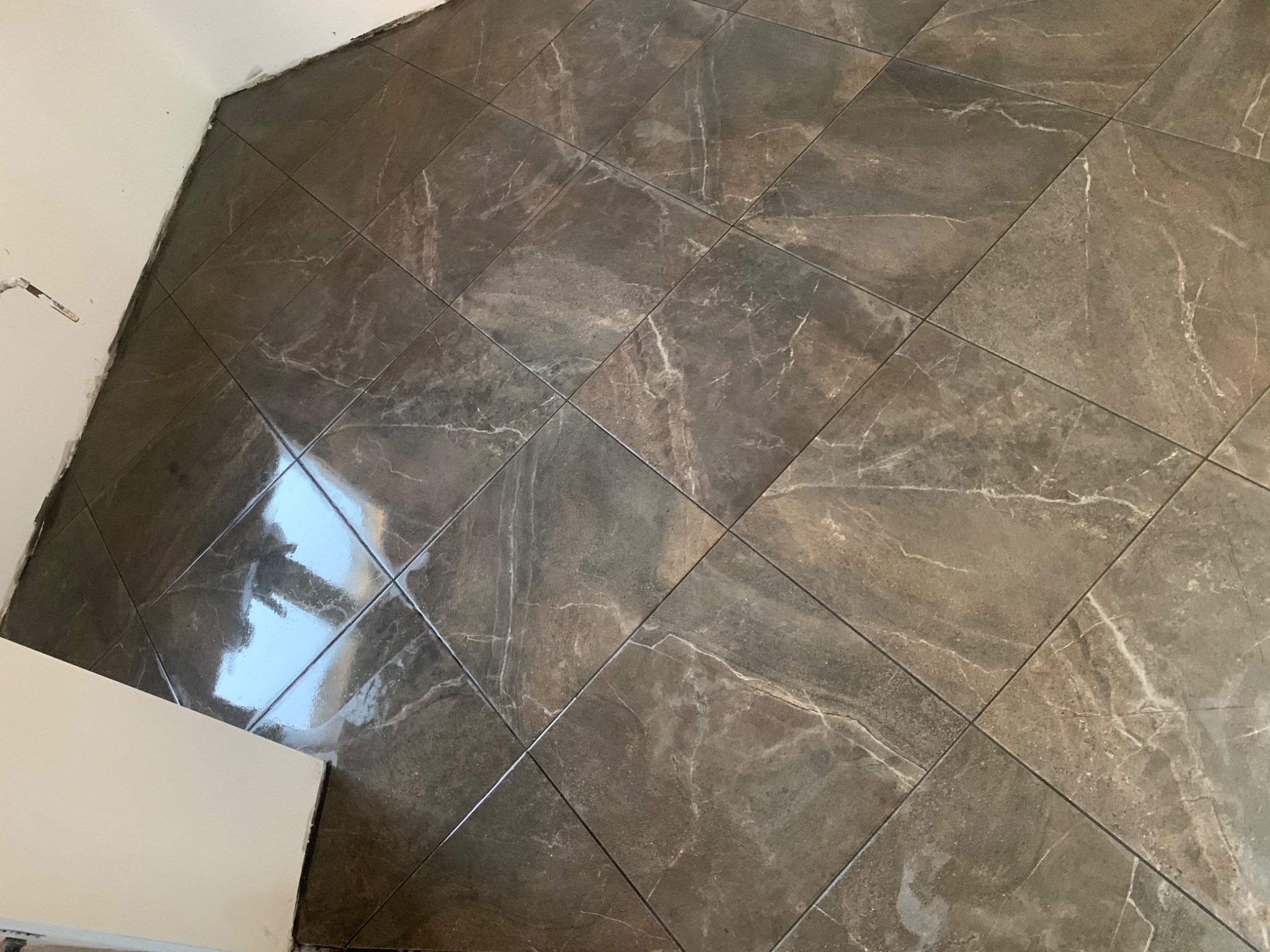 Ułożone brązowo-szare płytki podłogowe z efektem marmuru, widoczne niedociągnięcia przy ścianie, odbicie światła na powierzchni płytek.