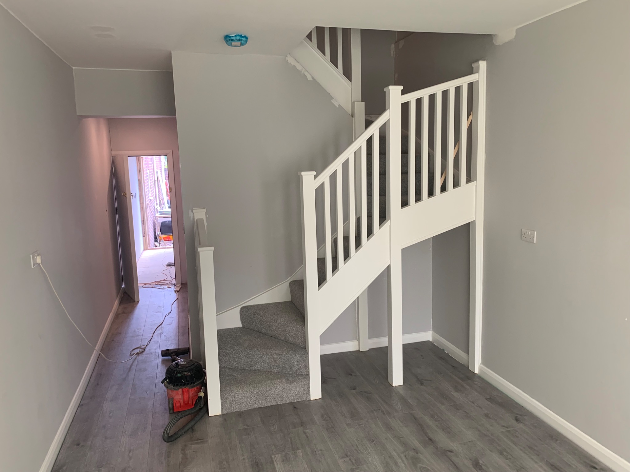 Pomieszczenie po remoncie: szare ściany, białe schody z szarym dywanem, szara podłoga, widoczny odkurzacz i kabel na podłodze.