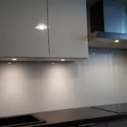 Szklany panel ścienny w kuchni, zamontowany między blatem a szafkami wiszącymi, podświetlony punktowymi lampami LED, z widocznym zlewem i płytą indukcyjną.