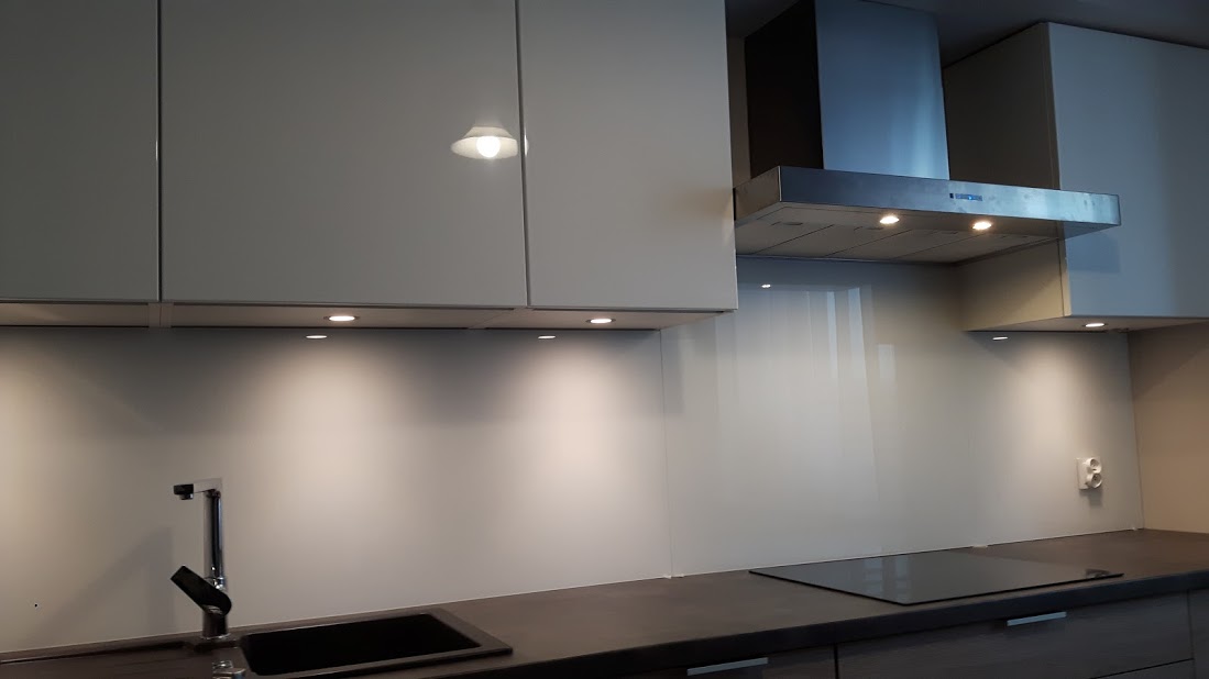 Szklany panel ścienny w kuchni, zamontowany między blatem a szafkami wiszącymi, podświetlony punktowymi lampami LED, z widocznym zlewem i płytą indukcyjną.