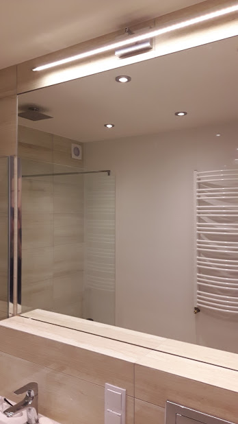 Duże lustro łazienkowe z oświetleniem LED, odbijające kabinę prysznicową i grzejnik drabinkowy. Widoczne fragmenty umywalki i baterii.