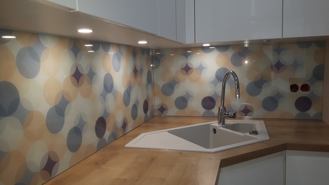 Szklany panel kuchenny z kolorowym motywem kół, oświetlenie punktowe pod szafkami, zlewozmywak narożny z baterią.