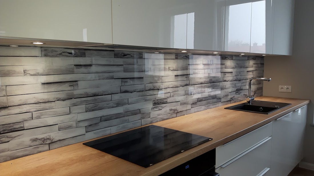 Szklany panel kuchenny z motywem szarej cegły, zamontowany między białymi szafkami a drewnianym blatem, z widoczną czarną płytą indukcyjną i zlewozmywakiem.