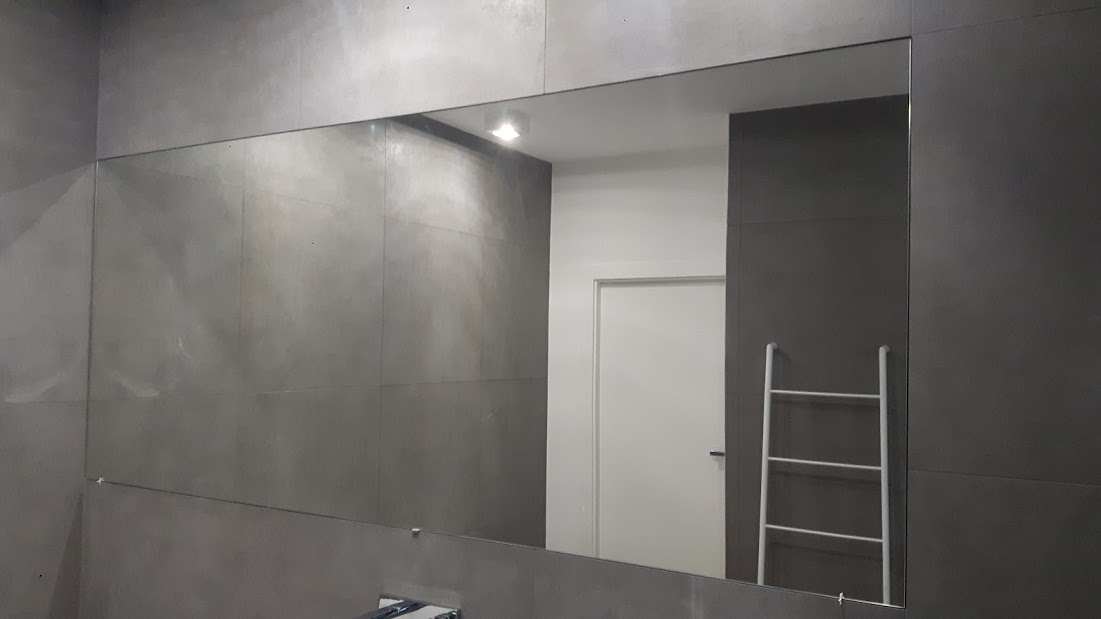 Prostokątne lustro wklejone w szarą ścianę łazienki, odbijające fragment białych drzwi i biały drabinkowy wieszak na ręczniki.