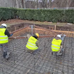 Trzech pracowników w odblaskowych kamizelkach montuje zbrojenie fundamentu, widoczne szalunki i wystające elementy instalacji.