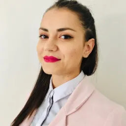 Portret uśmiechniętej kobiety w różowej marynarce i białej koszuli, z makijażem i ciemnymi włosami zaczesanymi do tyłu.