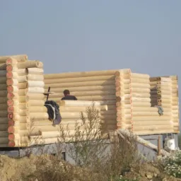 Budowa domu z bali w trakcie realizacji, widoczne oznaczenia na belkach i pracownik wewnątrz konstrukcji, na pierwszym planie ziemia i roślinność.