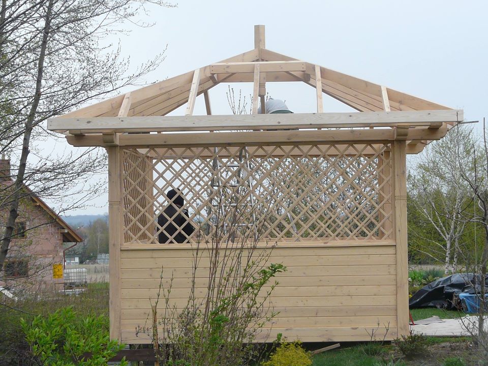 Konstrukcja altany ogrodowej z drewnianą kratownicą, widoczne elementy więźby dachowej w trakcie montażu, na drugim planie budynek mieszkalny.