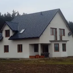 Nowo wybudowany dom z ciemną dachówką w otoczeniu lasu, widoczne okna z drewnianymi ramami i częściowo zagospodarowany teren wokół budynku.