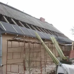 Dom jednorodzinny w trakcie prac dekarskich: częściowo zdjęte stare pokrycie, widoczna więźba, fragment dachu zabezpieczony czarną folią, rusztowania przy ścianie, przygotowane łaty dachowe...