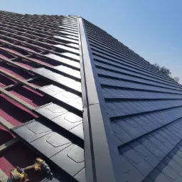 Montaż ciemnoszarej dachówki na drewnianej konstrukcji dachu, widoczna wkrętarka akumulatorowa, błękitne niebo w tle.