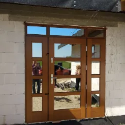 Brązowe, przeszklone wejście do budynku w trakcie budowy, z odbiciami osób i samochodu w szybach. Widoczne białe bloczki i fragment dachu.