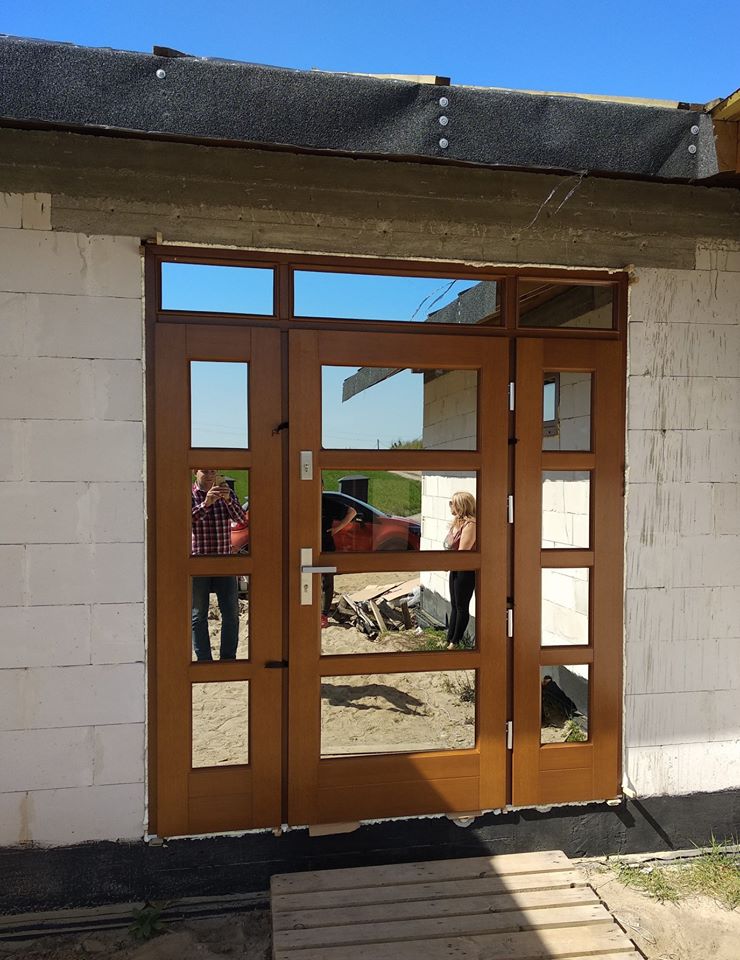 Brązowe, przeszklone wejście do budynku w trakcie budowy, z odbiciami osób i samochodu w szybach. Widoczne białe bloczki i fragment dachu.