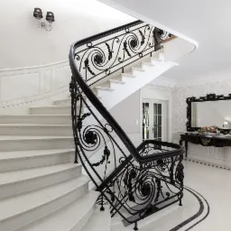 Eleganckie, kręte schody z białego marmuru z kutą, czarną balustradą o dekoracyjnych wzorach roślinnych, w przestronnym holu z lustrem w czarnej ramie i wzorzystą tapetą.