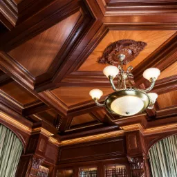 Bogato zdobiony, drewniany sufit kasetonowy z ornamentami i centralnie umieszczonym żyrandolem, widok z dołu na klasyczną biblioteczkę z książkami i delikatną zasłoną w tle.