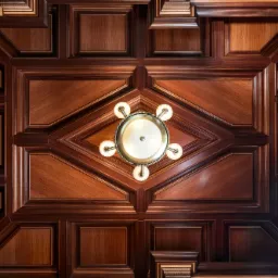 Kasetonowy sufit z ciemnego drewna z geometrycznymi wzorami i centralnie umieszczonym żyrandolem z pięcioma szklanymi kloszami, widoczne logo firmy w prawym górnym rogu.