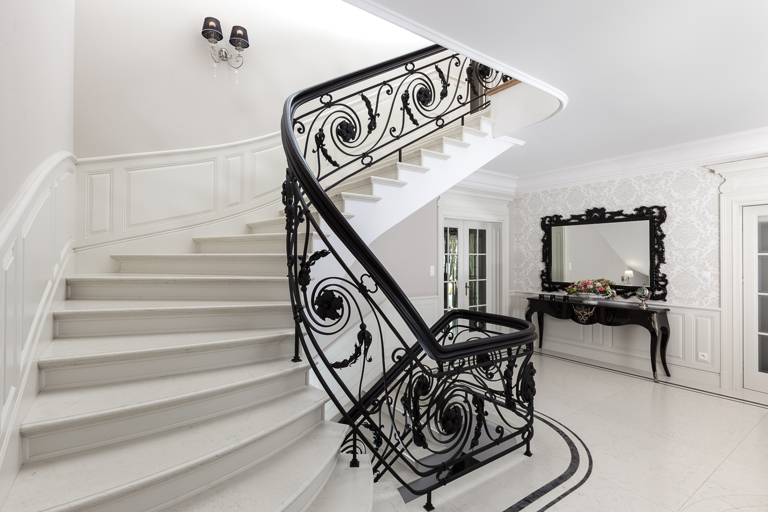 Eleganckie, kręte schody z białego marmuru z kutą, czarną balustradą o dekoracyjnych wzorach roślinnych, w przestronnym holu z lustrem w czarnej ramie i wzorzystą tapetą.