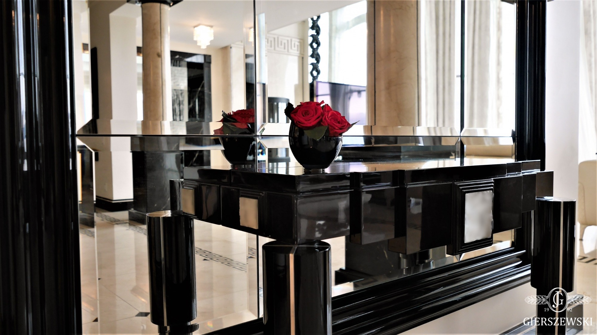 Błyszczący, czarny stół w stylu art deco z wazonem czerwonych róż na blacie, odbijający się w lustrzanej ścianie w eleganckim wnętrzu z marmurową podłogą i kolumnami.