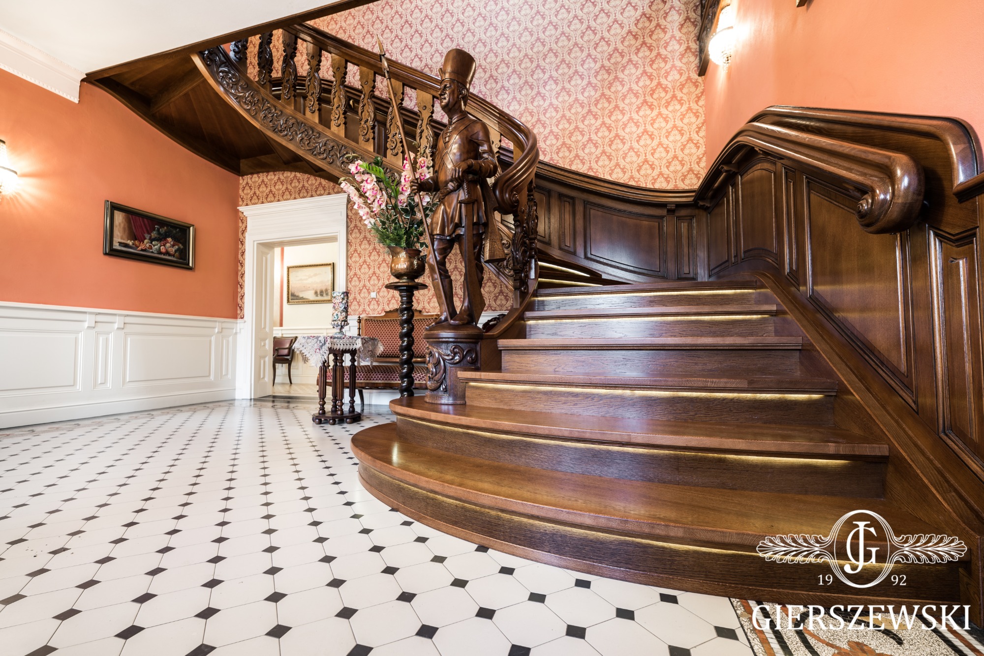 Eleganckie, kręte schody drewniane z podświetleniem LED, zdobione rzeźbioną figurą żołnierza, w holu z wzorzystą podłogą i tapetą.