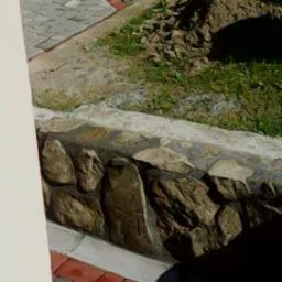 Fragment murku oporowego z nieregularnych kamieni, połączonych jasną zaprawą, z widocznym trawnikiem i fragmentem chodnika z kostki w tle, ujęcie z niskiej perspektywy.