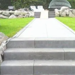 Schody zewnętrzne z szarych, prostokątnych płyt kamiennych prowadzące do ścieżki i tarasu. W tle widoczny trawnik, kamienny murek i leżaki.