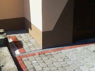 Ułożona kostka brukowa w odcieniach szarości z czerwonym obramowaniem przy narożniku budynku, widoczna kratka odpływowa, niedokończone obrzeże po lewej stronie.