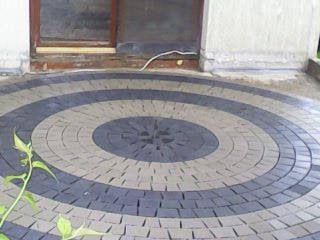 Okrągły wzór z kostki brukowej w odcieniach szarości i beżu, ułożony na tarasie przed wejściem do budynku. Widoczny fragment ściany i rośliny w lewym dolnym rogu.