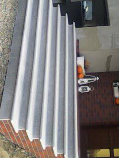 Zewnętrzne schody z szarego kamienia z białymi krawędziami, prowadzące do drzwi wejściowych z ceglaną ścianą, widoczne owoce i akcesoria na ścianie.