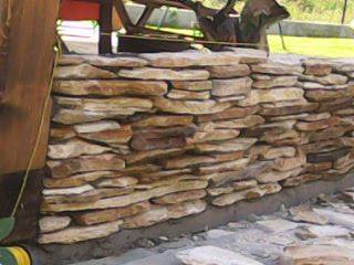 Fragment muru z nieregularnych, płaskich kamieni piaskowca w odcieniach beżu i brązu, ułożonych warstwowo, z widocznymi spoinami. Mur opiera się na betonowej podstawie, w tle zieleń trawnika i drzewa.