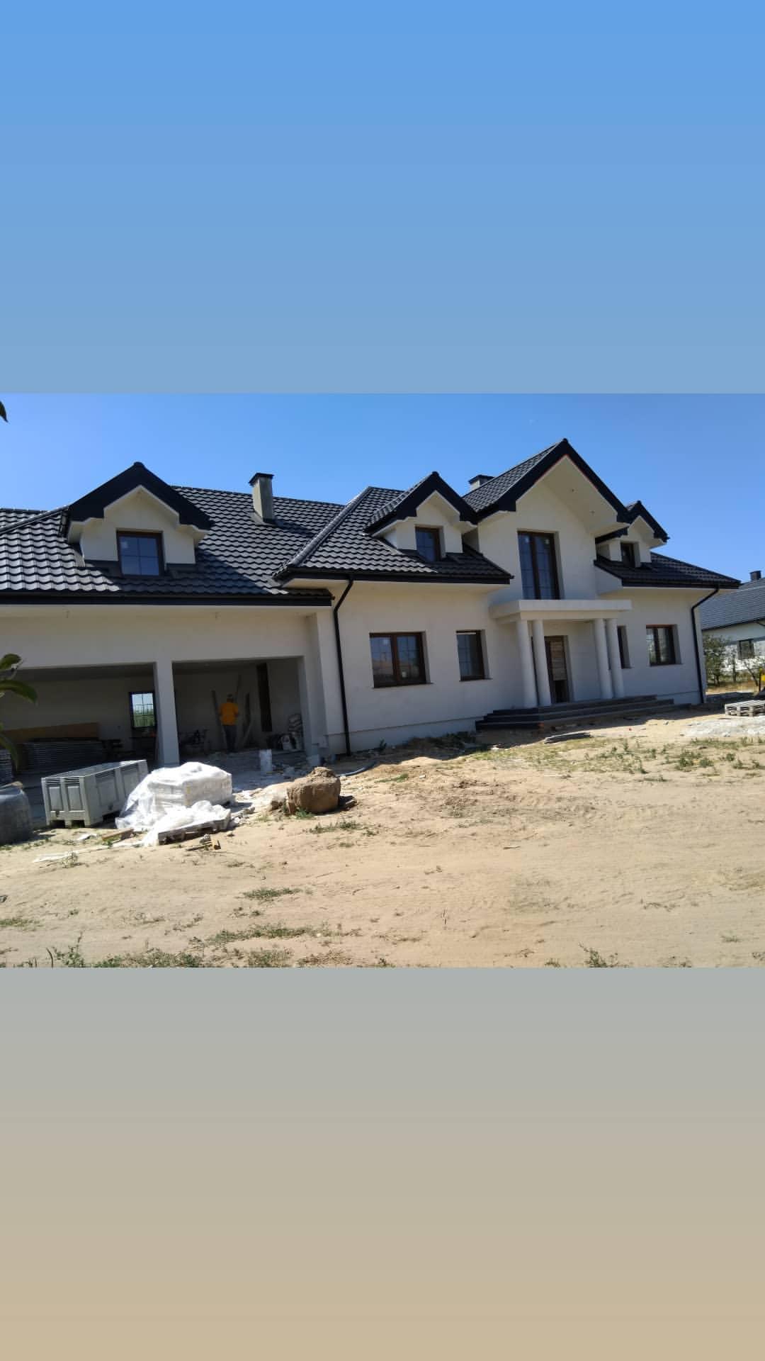 Nowoczesny dom jednorodzinny z białymi ścianami i czarnym dachem, w trakcie wykańczania elewacji, widoczny otwarty garaż z elementami budowlanymi w środku.