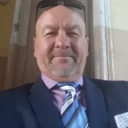 Portret uśmiechniętego mężczyzny w garniturze, z okularami na głowie, na tle kolumn w odcieniach beżu i brązu.